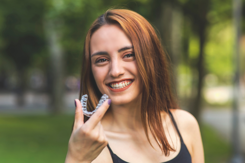 A cheerful young woman with long auburn hair smiles confidently while holding a clear Invisalign aligner in her hand at a park – Invisalign