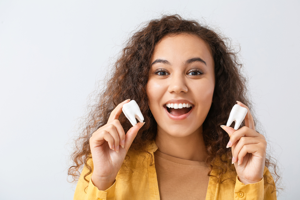 A happy young woman with curly hair holds two oversized tooth models in each hand, smiling widely in a playful and educational dental context – Extractions