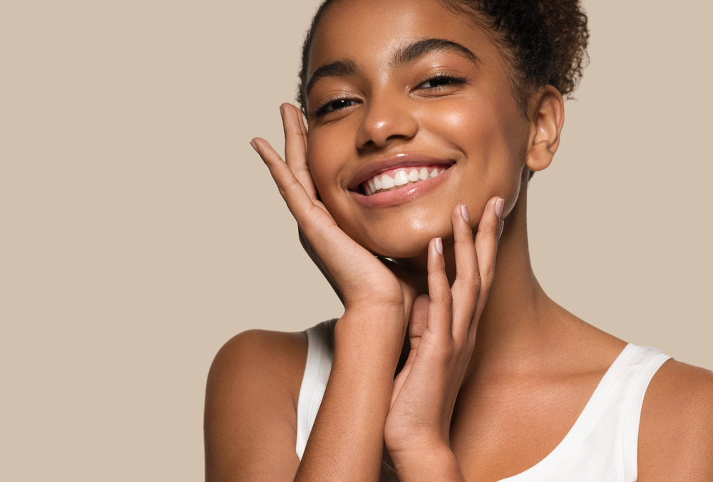 A woman with flawless skin and a radiant, even smile poses with her hands framing her face, showcasing the natural beauty of porcelain veneers - Bellevue Porcelain Veneers 