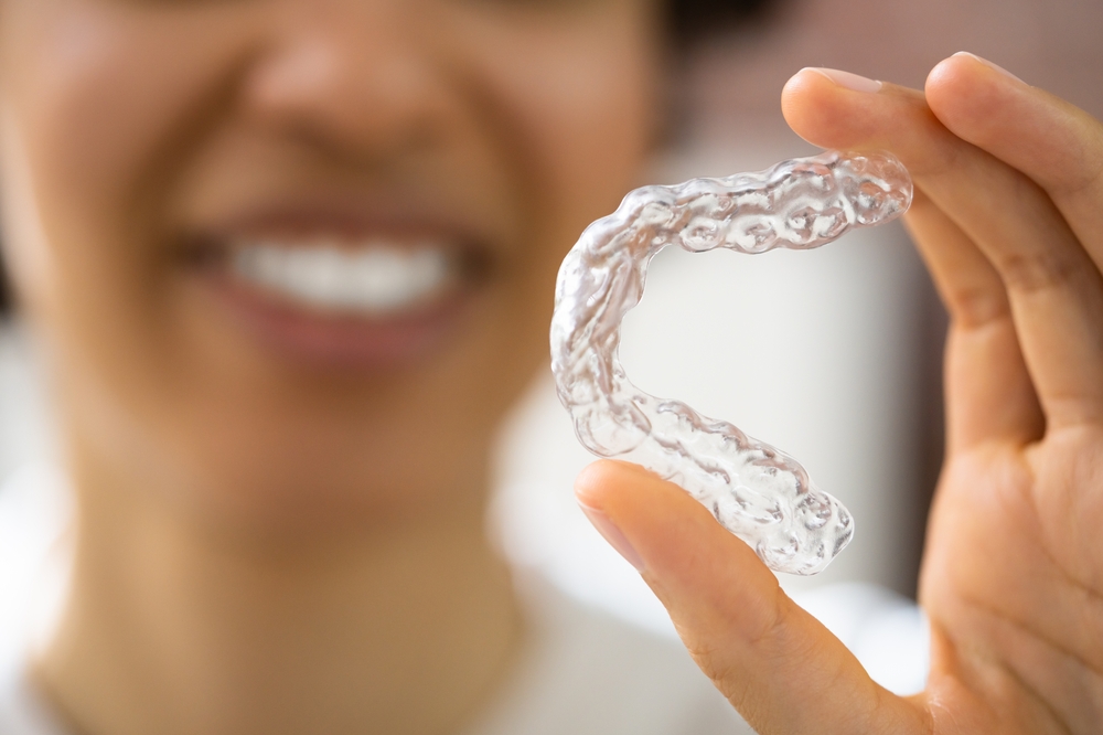 A woman smiles in the background while holding a transparent Invisalign aligner between her fingers in the foreground – Invisalign