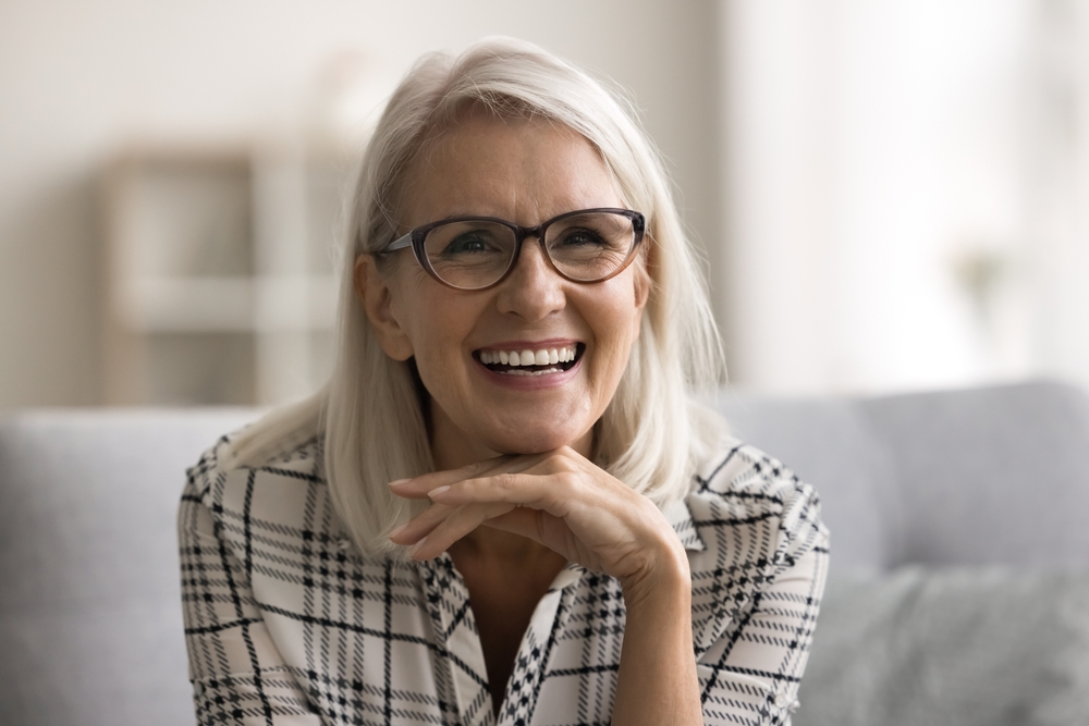 Happy Senior Woman with Glasses Smiling on Couch – Dentures A cheerful senior woman with white hair and stylish glasses smiles warmly while relaxing on a couch, showcasing a confident and healthy smile – Dentures