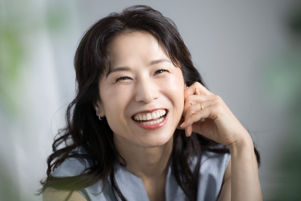 Confident Middle-Aged Woman Smiling with Healthy Teeth – Dentures A joyful middle-aged woman with long dark hair and radiant white teeth smiles confidently while resting her hand on her cheek, highlighting dental wellness – Dentures