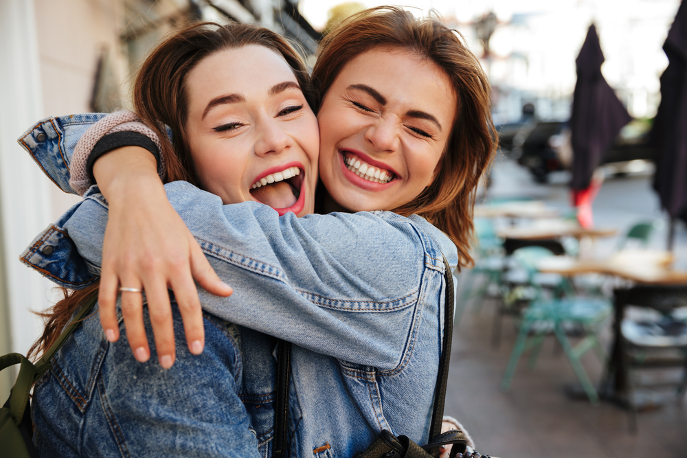 Laughing Friends with Bright Smiles - Dental Crowns Bellevue Two young women joyfully hugging on a city street, both showing off radiant smiles that reflect the confidence and beauty achieved with dental crowns - Dental Crowns Bellevue