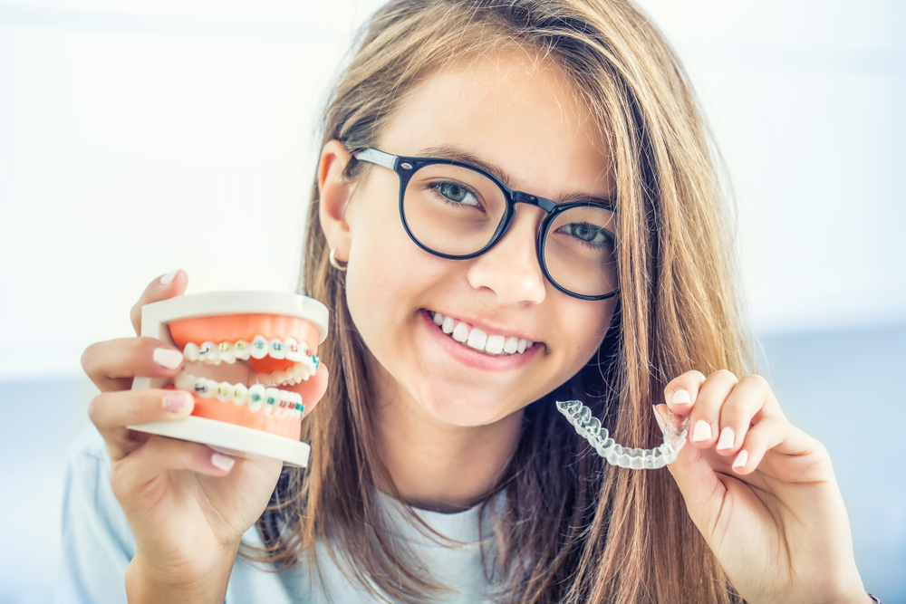 A cheerful teenage girl wearing glasses holds a dental model with traditional braces in one hand and a clear aligner in the other, showcasing orthodontic treatment options – Dentures