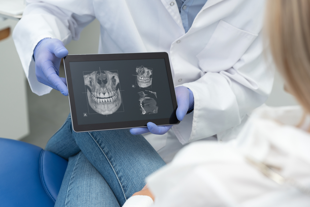 Dental Consultation Showing 3D Jaw X-Ray on Tablet - Dental X-Rays A dentist wearing gloves and a white coat shows a patient detailed 3D dental X-ray images on a tablet, highlighting jaw and teeth structure during a consultation – Dental X-Rays