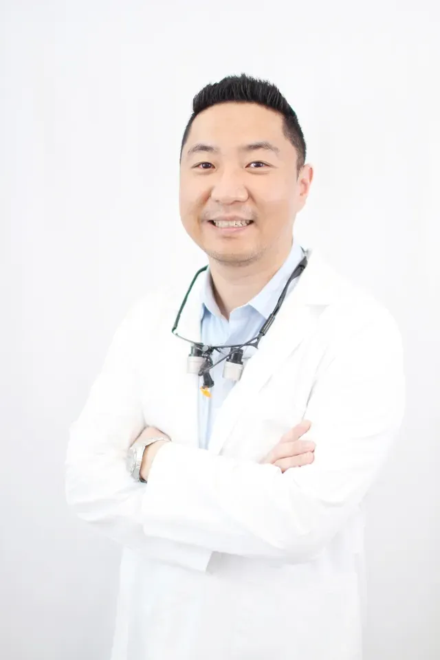 Dr. Alex Shin, DMD in a white coat, wearing dental loupes around his neck, smiles with arms crossed against a plain white background - Newport Dental Bellevue WA Dr. Alex Shin, DMD in a white coat, wearing dental loupes around his neck, smiles with arms crossed against a plain white background - Newport Dental Bellevue WA