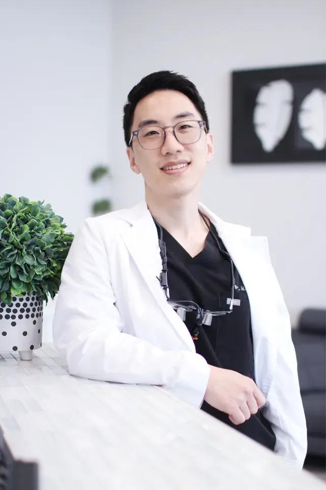 Dr. Brian Han, DMD in a white coat and glasses smiles confidently while leaning on a counter in a modern, professional office with minimalist decor - Newport Dental Bellevue WA Dr. Brian Han, DMD in a white coat and glasses smiles confidently while leaning on a counter in a modern, professional office with minimalist decor - Newport Dental Bellevue WA