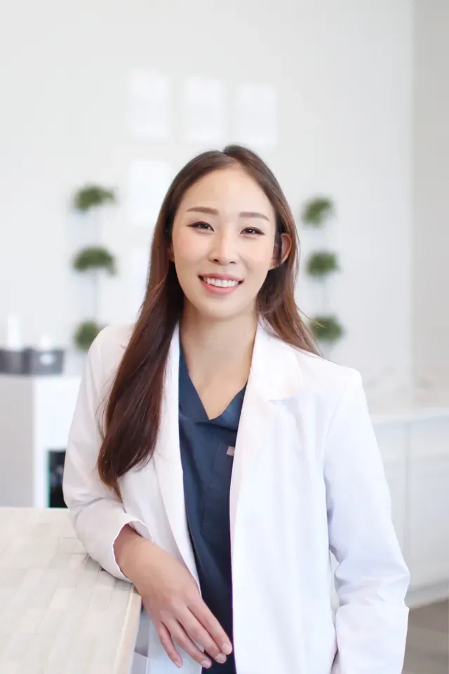 Dr. Sarah Park, DDS in a white coat stands in a clean, bright office environment with decorative greenery in the background - Newport Dental Bellevue WA Dr. Sarah Park, DDS in a white coat stands in a clean, bright office environment with decorative greenery in the background - Newport Dental Bellevue WA