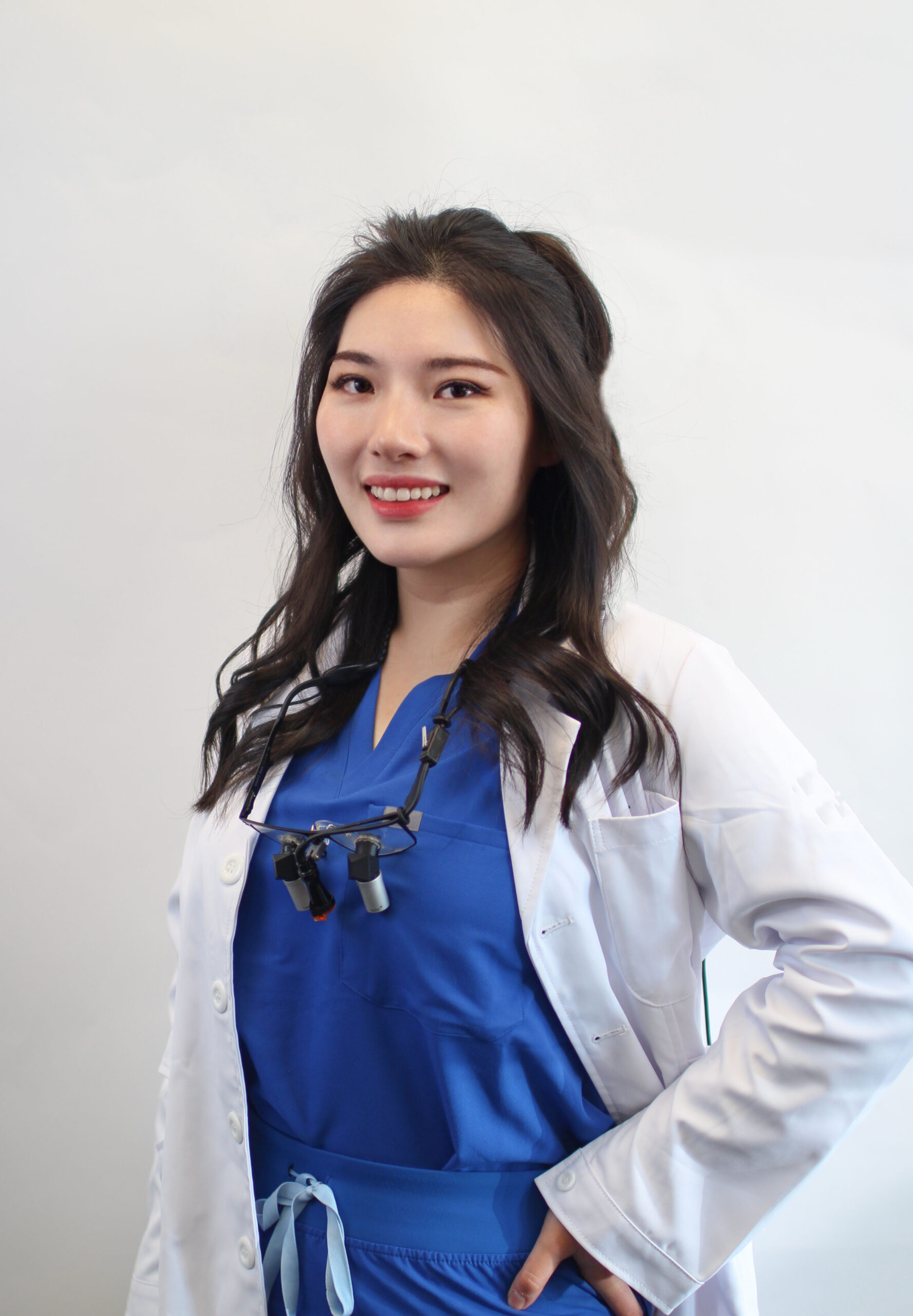 Dr. Yongxi Liang, DMD in a white coat and pink scrubs, wearing dental loupes around her neck, smiles warmly in a clinic setting - Newport Dental Bellevue WA
