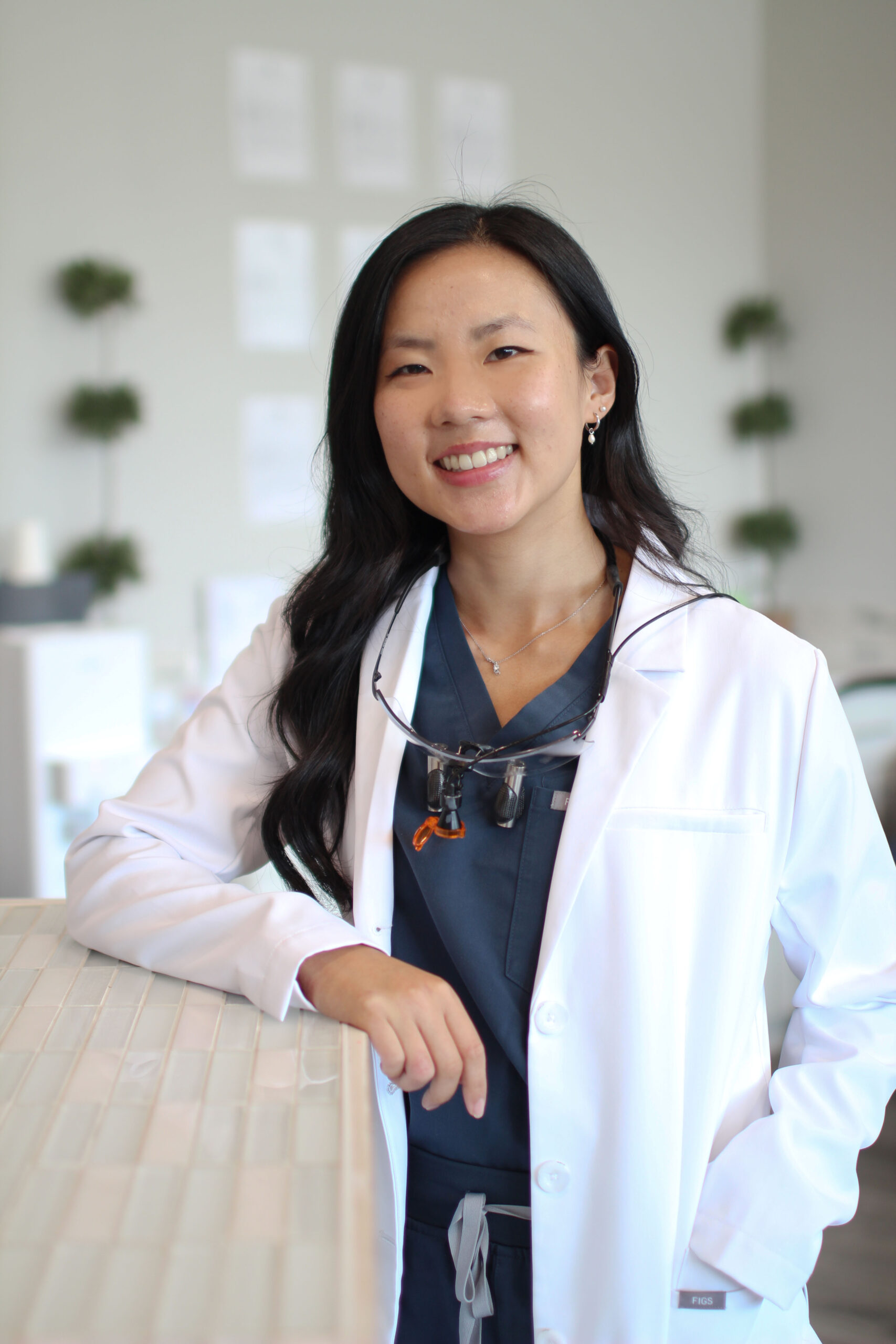 Dr. Brian Han, DMD in a white coat and glasses smiles confidently while leaning on a counter in a modern, professional office with minimalist decor - Newport Dental Bellevue WA