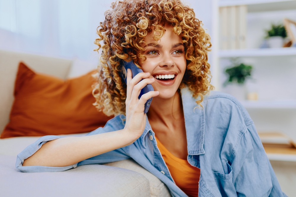 A woman with curly hair smiles brightly while talking on a phone, sitting in a cozy living room - Newport Dental Bellevue WA A woman with curly hair smiles brightly while talking on a phone, sitting in a cozy living room - Newport Dental Bellevue WA