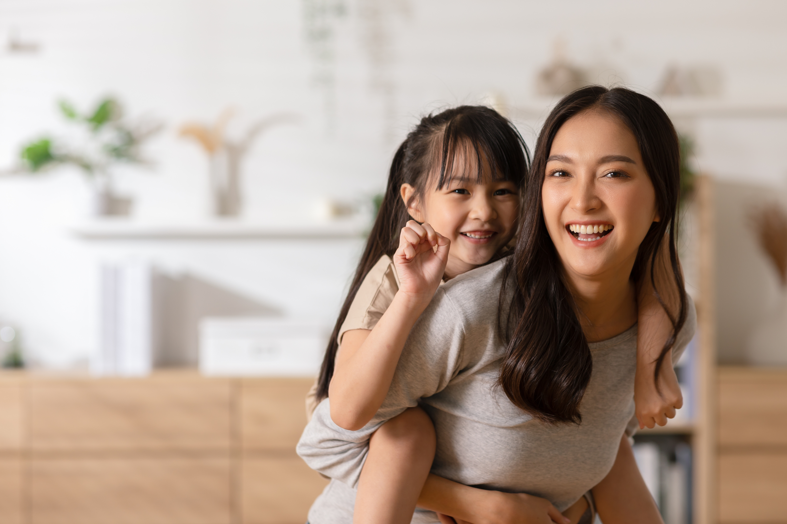 Happy Mother and Daughter Smiling at Home - Dental Fillings Joyful mother giving piggyback ride to daughter, both smiling brightly in a cozy home environment - Dental Fillings