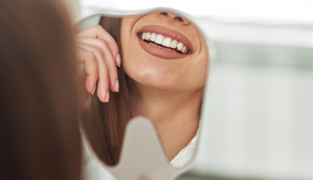 Confident Smile After Cleaning and Exams Close-up of a woman smiling at her reflection in a tooth-shaped mirror after a professional dental cleaning and exam