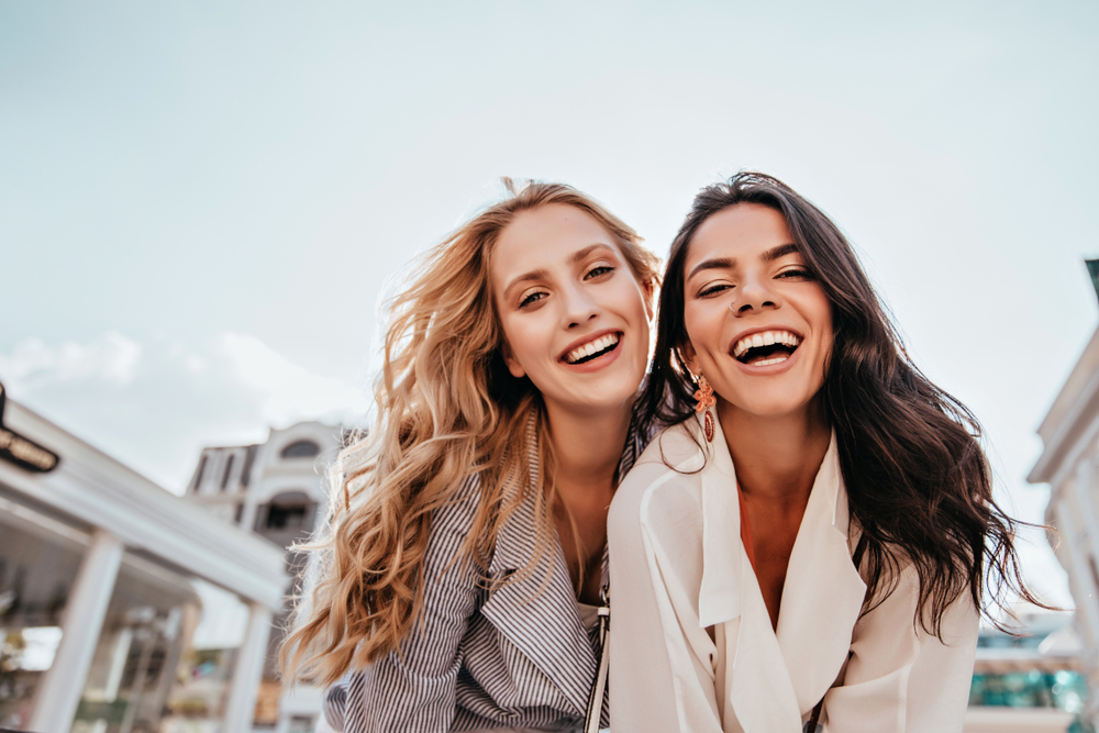 Two Smiling Women Outdoors with Bright, Healthy Teeth – General Dentistry Two joyful young women with radiant smiles and healthy teeth pose together outdoors in bright natural light, showcasing results of routine dental care – General Dentistry