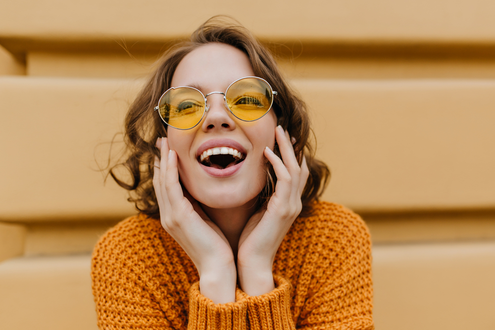 Cheerful Young Woman in Sunglasses Smiling with Open Mouth – General Dentistry A happy young woman wearing yellow-tinted sunglasses and an orange sweater smiles brightly with her mouth open, showing healthy teeth – General Dentistry