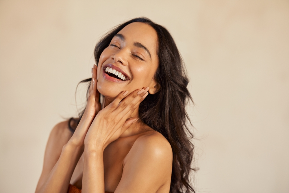 Radiant Laughing Woman - Bellevue Cosmetic Dentistry A joyful woman with glowing skin and a flawless smile touches her face while laughing, representing confidence after cosmetic dental enhancements - Bellevue Cosmetic Dentistry