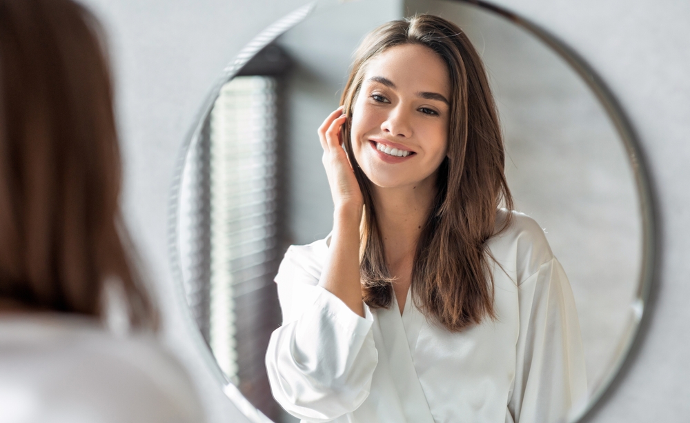 A young woman in a white robe smiles confidently while looking at herself in a round mirror, suggesting satisfaction after a successful dental treatment – Extractions