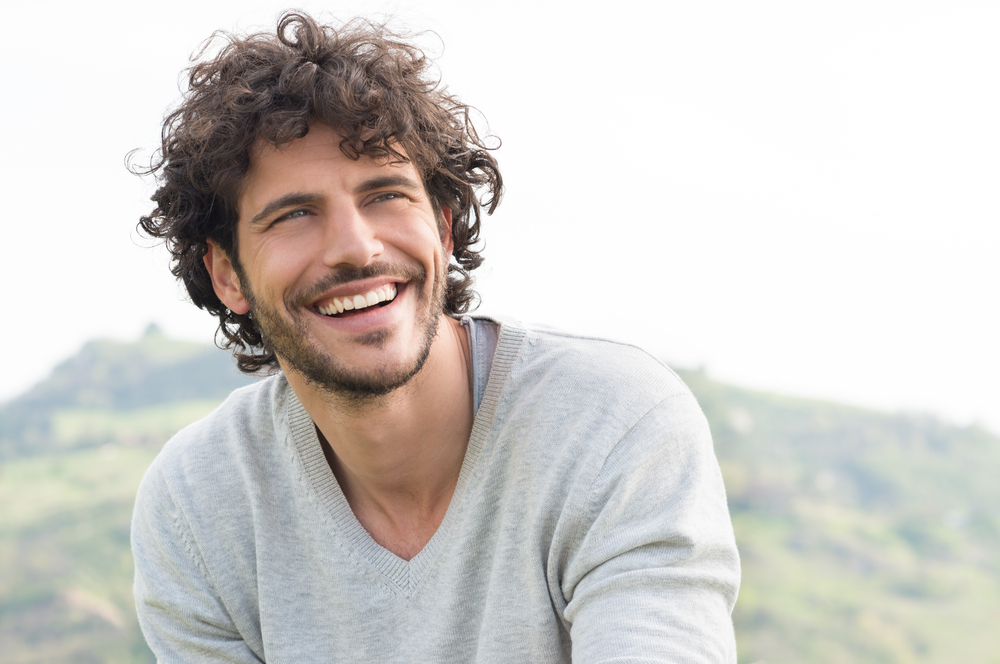 Bright Outdoor Smile - Teeth Whitening Bellevue A smiling man with curly hair outdoors in a light sweater, displaying a naturally bright smile achieved through professional whitening - Teeth Whitening Bellevue