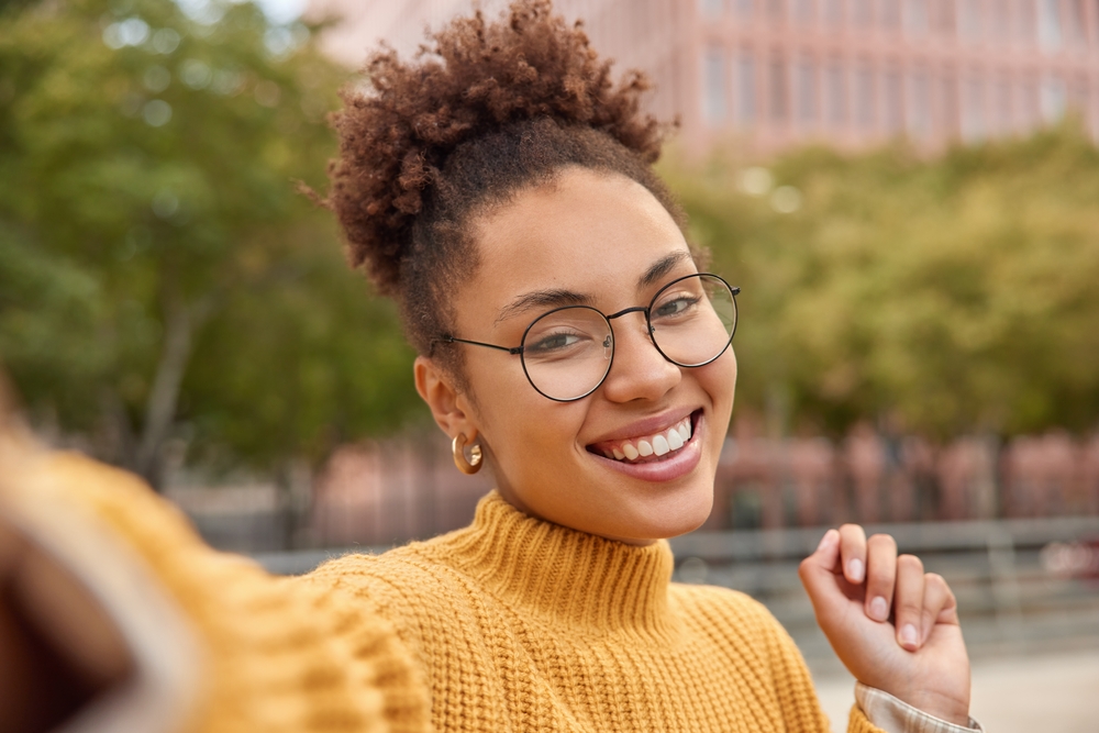 Confident Outdoor Smile in Glasses - Dental Fillings Young woman wearing glasses and a yellow sweater smiling outdoors with perfect teeth - Dental Fillings