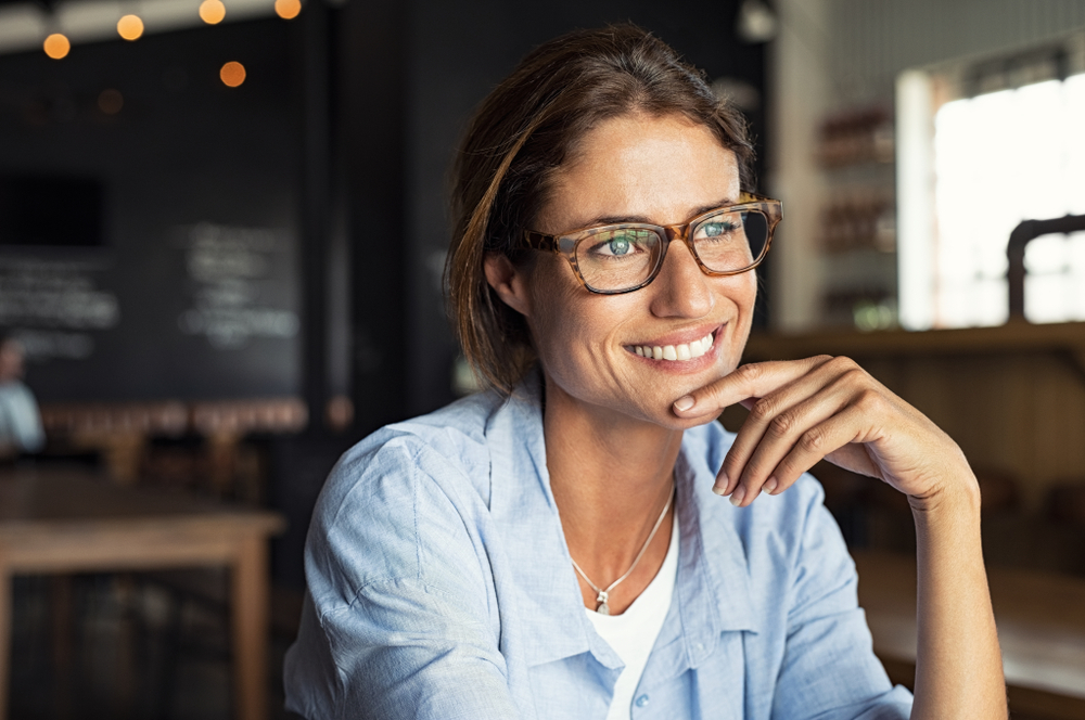 Stylish middle-aged woman with glasses smiling thoughtfully in a café, her radiant teeth reflecting the benefits of porcelain veneers - Bellevue Porcelain Veneers