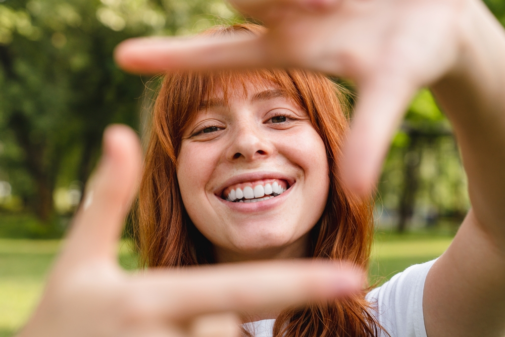 Joyful Smile After Treatment - Dental Implants Red-haired woman smiling brightly and framing her face with hands outdoors, symbolizing confidence after dental implants - Dental Implants