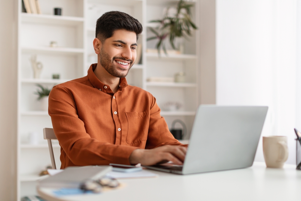 A man in a casual orange shirt smiles while working on a laptop at a bright, modern desk - Newport Dental Bellevue WA A man in a casual orange shirt smiles while working on a laptop at a bright, modern desk - Newport Dental Bellevue WA