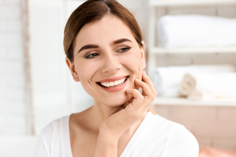 A beautiful woman in a white top gently touches her cheek while smiling, highlighting smooth, white teeth often achieved with dental veneers – Veneers