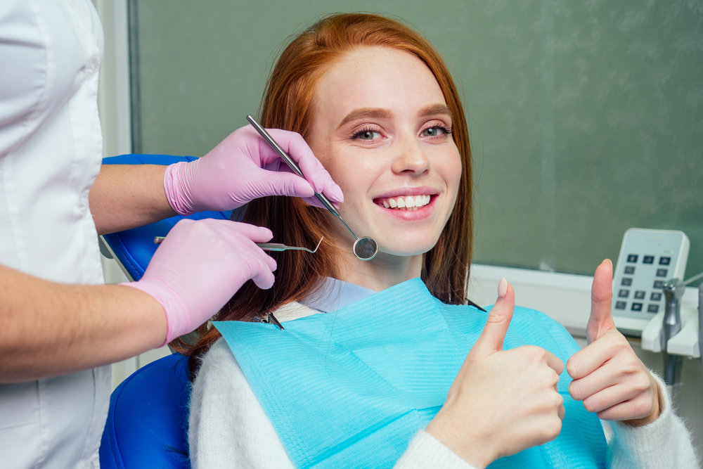 Confident Smile After Treatment - Cosmetic Dentistry Happy red-haired woman giving thumbs up during dental exam with tools in mouth - Cosmetic Dentistry