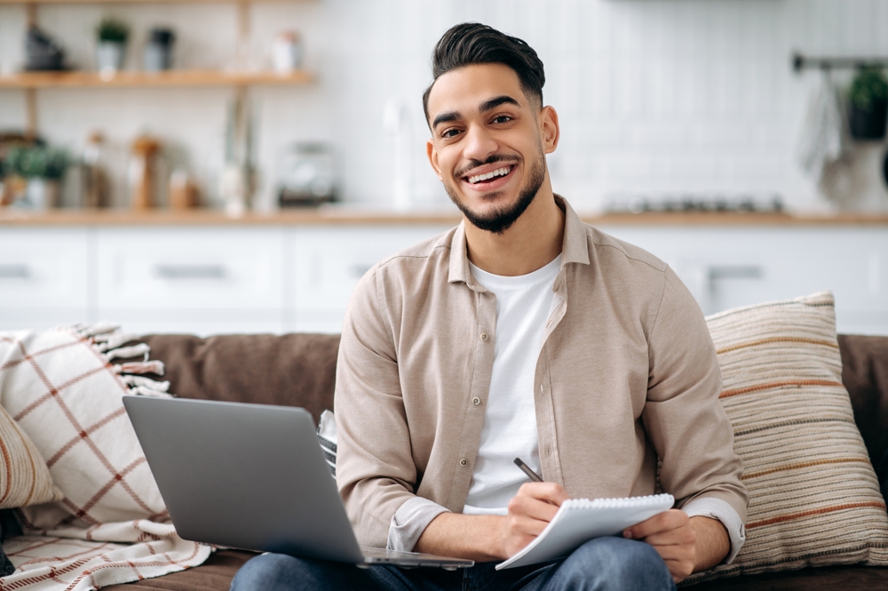 A smiling man sits on a couch with a laptop and notepad, appearing relaxed and productive in a cozy home setting - Newport Dental Bellevue WA A smiling man sits on a couch with a laptop and notepad, appearing relaxed and productive in a cozy home setting - Newport Dental Bellevue WA