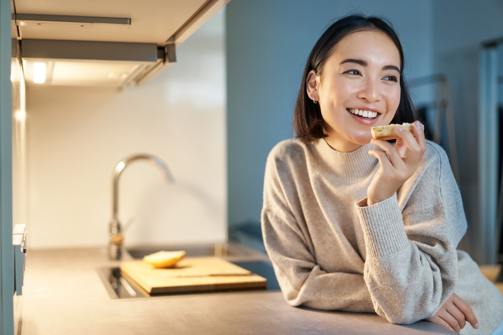 Happy Woman Enjoying Food at Home with a Healthy Smile – Dental Insurance A cheerful young woman leans on a kitchen counter while eating and smiling confidently, representing the benefits of dental insurance for maintaining oral health – Dental Insurance