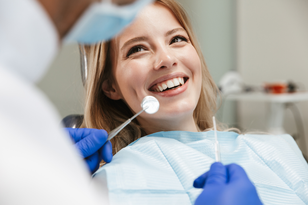 Smiling Woman in Dental Chair During Exam with Mirror Tool – Root Canals A happy female patient sits in a dental chair, smiling as a gloved dentist examines her teeth with a mirror and probe during a root canal consultation – Root Canals