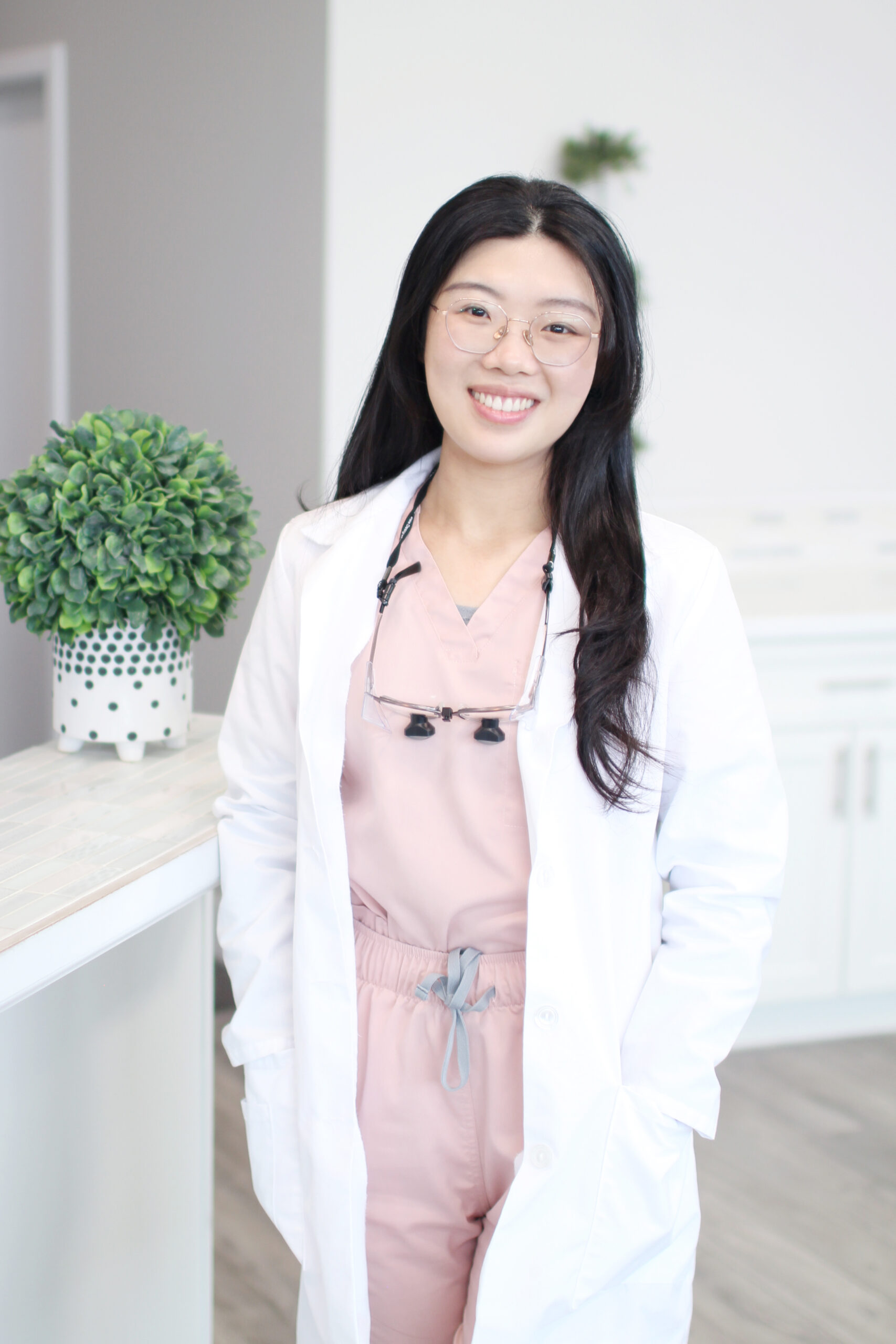Dr. Yongxi Liang, DMD in a white coat and pink scrubs, wearing dental loupes around her neck, smiles warmly in a clinic setting - Newport Dental Bellevue WA Dr. Yongxi Liang, DMD in a white coat and pink scrubs, wearing dental loupes around her neck, smiles warmly in a clinic setting - Newport Dental Bellevue WA